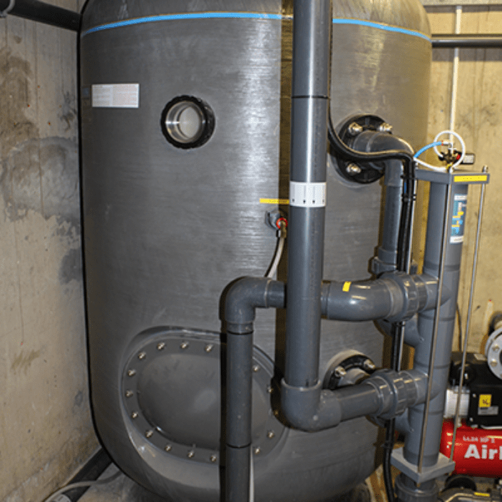 Large cylindrical black water tank stands connected to several grey PVC pipes and valves, circulating and filtering fluid inside a concrete utility room with a floor-mounted compressor and exposed wiring.