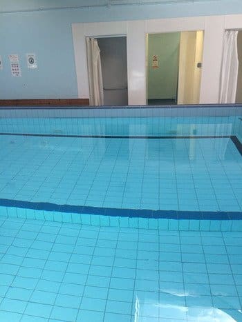Indoor tiled pool lies calm, reflecting lights; a shallow ledge and dark lane cross the blue water, with changing-room doorways and curtained alcoves on a pale wall. No legible text.