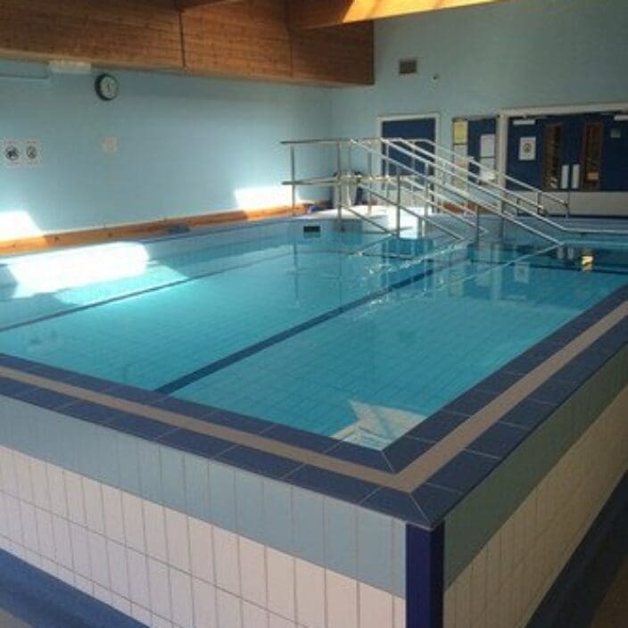 Indoor rectangular swimming pool sits filled with clear water reflecting a skylight, stainless steel handrails and steps lead into the pool, tiled blue deck and pale blue walls under wooden-beamed ceiling.