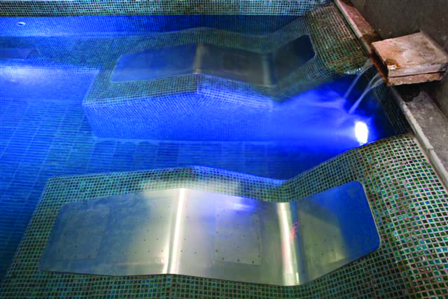 Two stainless-steel lounge panels lie submerged as water pours from a small spout and blue LED lights illuminate the mosaic-tiled pool/spa edge.