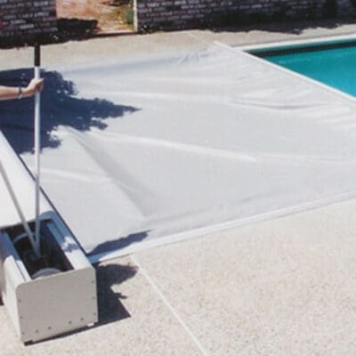 Pool cover being pulled across a residential swimming pool while a woman operates a manual reel on the concrete pool deck.