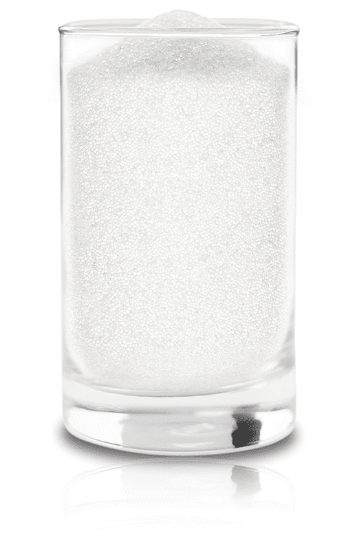 Clear cylindrical glass holds densely packed fine white granules to the rim, resting on a glossy white surface with a soft reflection, photographed against a seamless white background.