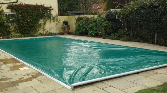 Rectangular outdoor swimming pool covered with a green protective tarp, sitting on a stone-paved patio, bordered by a wall with climbing plants and potted shrubs, dense hedges in the background.