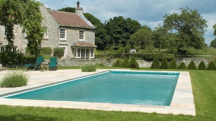 A rectangular turquoise swimming pool glistens, set in a stone patio with two green lounge chairs; a rustic stone country house and trimmed hedges and trees fill the surrounding lawn.