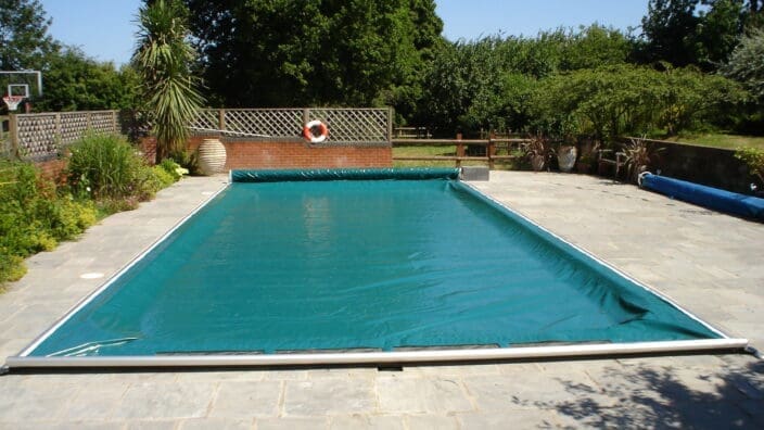 Rectangular swimming pool covered by a teal pool cover on a stone patio, surrounded by plants, trees, a lattice fence with a life preserver, under a clear blue sky.