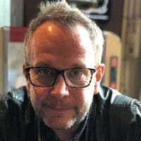 Person with short gray hair and glasses leans forward, looking directly at the camera; light stubble and a leather jacket suggest a casual indoor cafe or bar setting with blurred background.