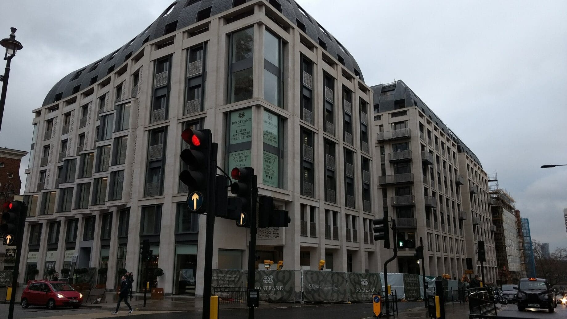 Large modern stone-and-glass building dominates an urban intersection; traffic lights show red while pedestrians cross and cars wait on wet pavement beneath an overcast sky. Text visible: "190 STRAND" "LUXURY APARTMENTS AVAILABLE NOW" "ST EDWARD" "LOOK LEFT".