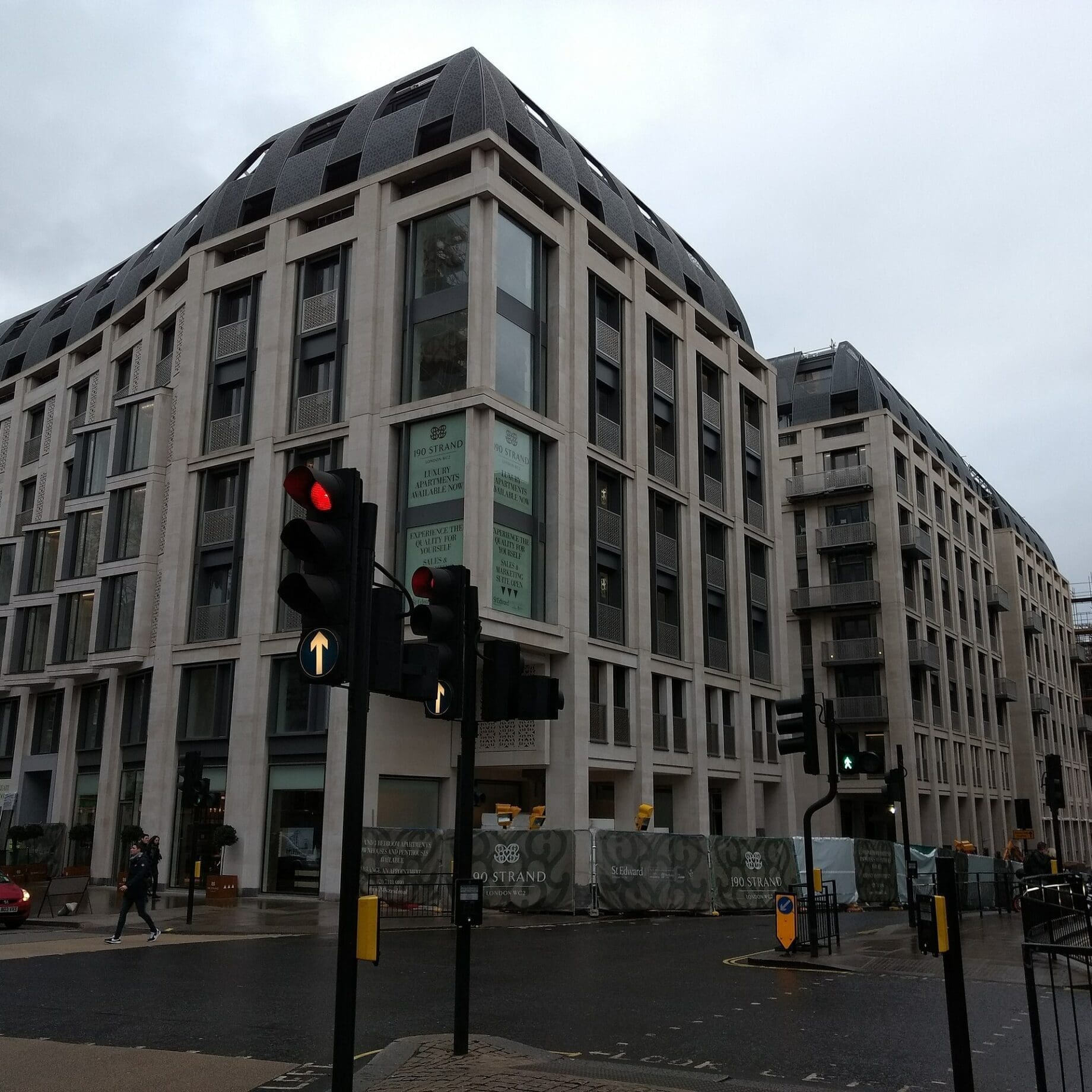 Large modern stone-and-glass building dominates an urban intersection; traffic lights show red while pedestrians cross and cars wait on wet pavement beneath an overcast sky. Text visible: "190 STRAND" "LUXURY APARTMENTS AVAILABLE NOW" "ST EDWARD" "LOOK LEFT".