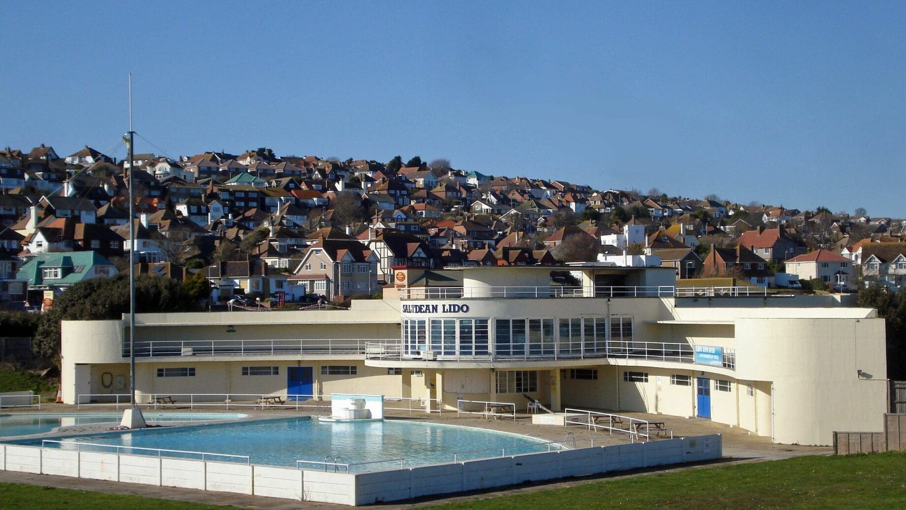 A cream lido building labeled SALTDEAN LIDO fronts a calm outdoor pool with no swimmers, surrounded by grassy lawns and a hillside of packed houses under a clear blue sky.