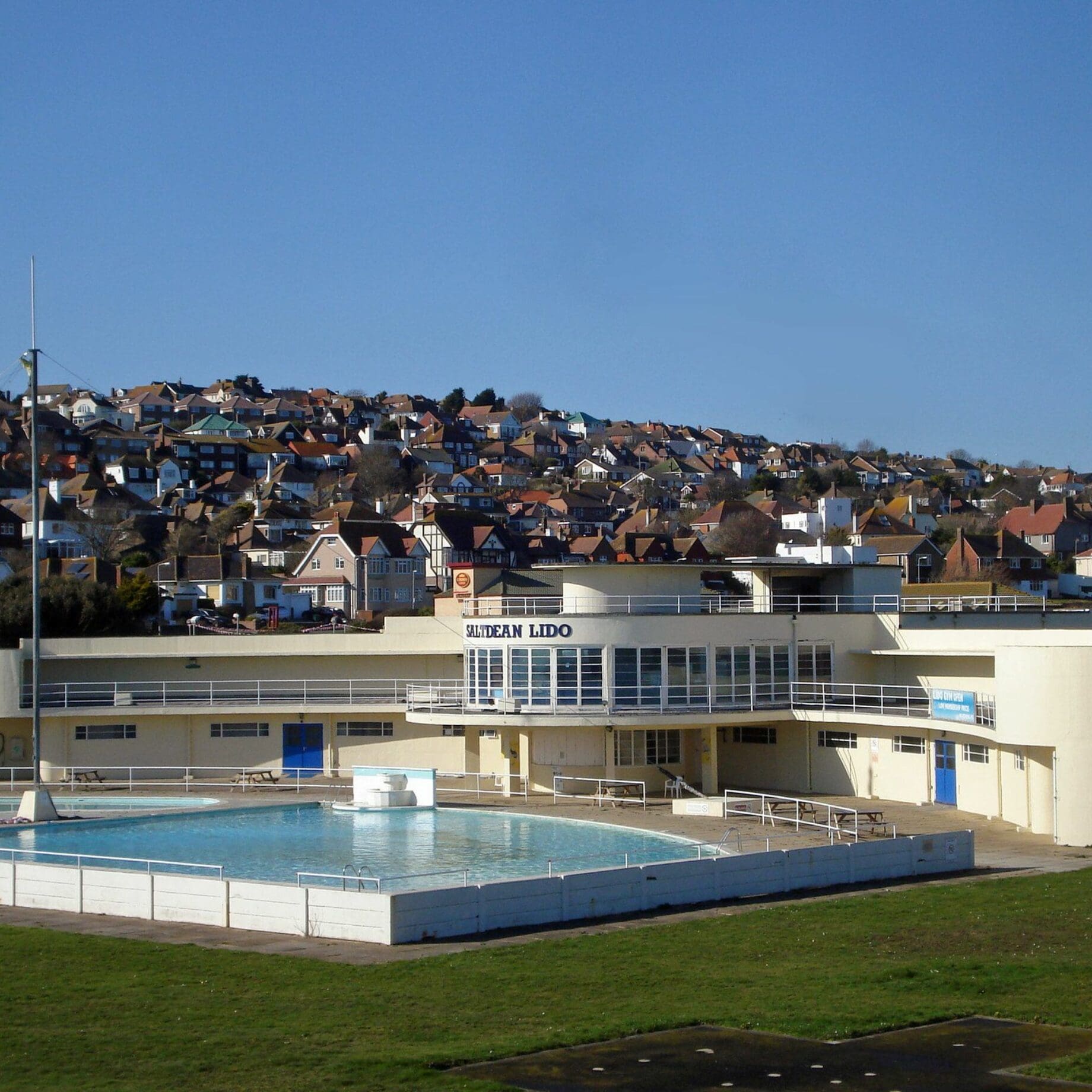 A cream lido building labeled SALTDEAN LIDO fronts a calm outdoor pool with no swimmers, surrounded by grassy lawns and a hillside of packed houses under a clear blue sky.