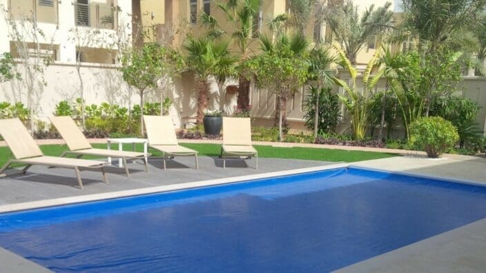 A rectangular blue-covered swimming pool sits still, bordered by lounge chairs and a small table on a wooden deck, with palm trees, shrubs and beige modern villas behind a low wall.