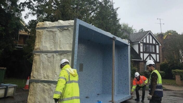 Large blue-tiled pool section being guided by three workers in high-visibility jackets and hard hats while suspended by chains on a wet suburban street with a Tudor-style house.