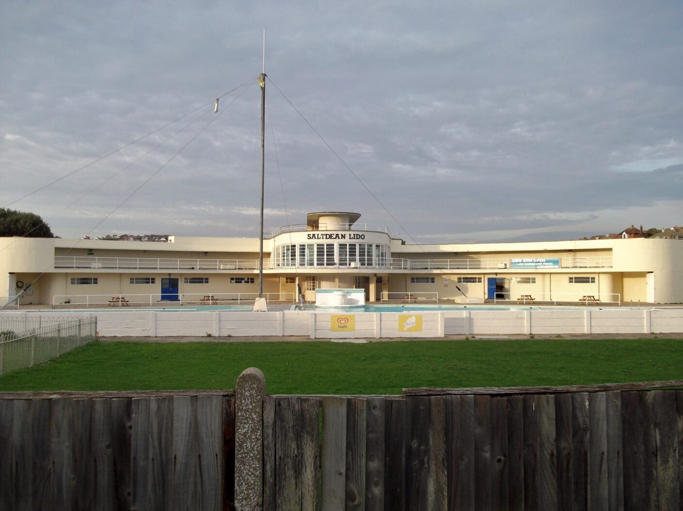 Saltdean Lido building overlooks an empty outdoor pool and picnic benches, set behind a grassy lawn and wooden fence under a cloudy sky. Text: SALTDEAN LIDO; Walls.