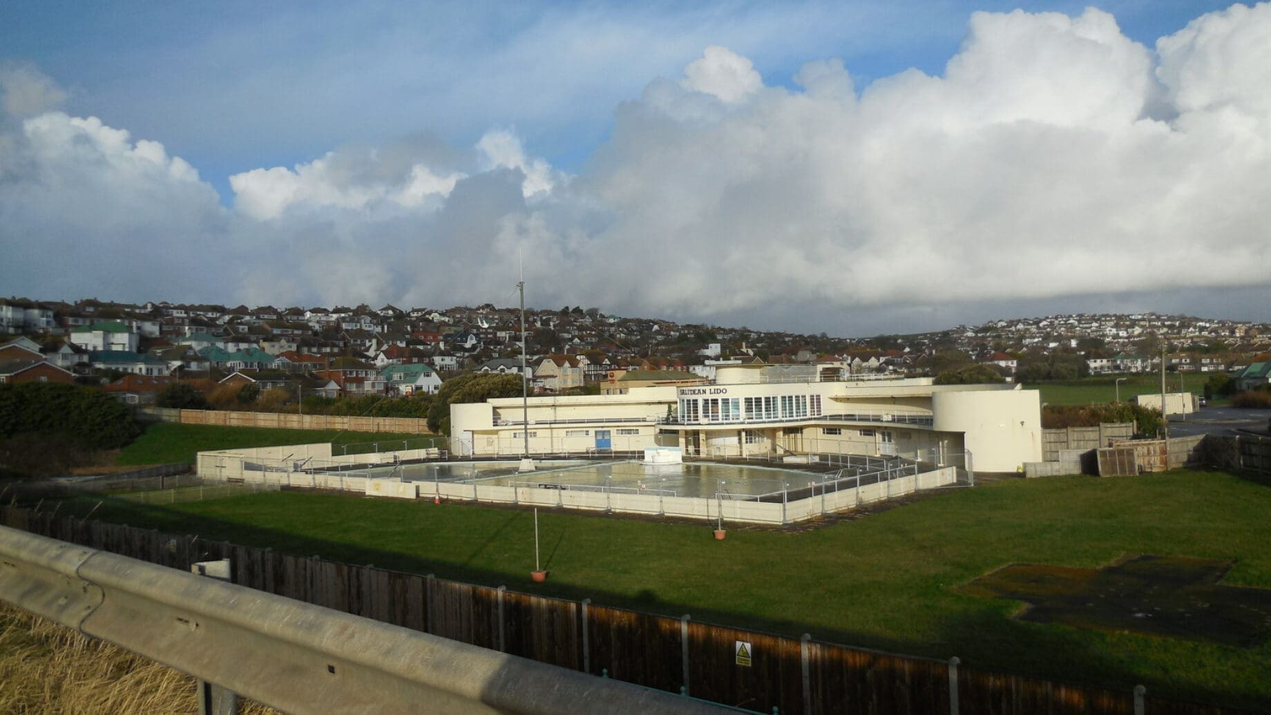 A pale lido building labeled SALTDEAN LIDO sits beside fenced empty outdoor pools on a grassy slope, with terraced houses on the hill and a cloud-filled blue sky.