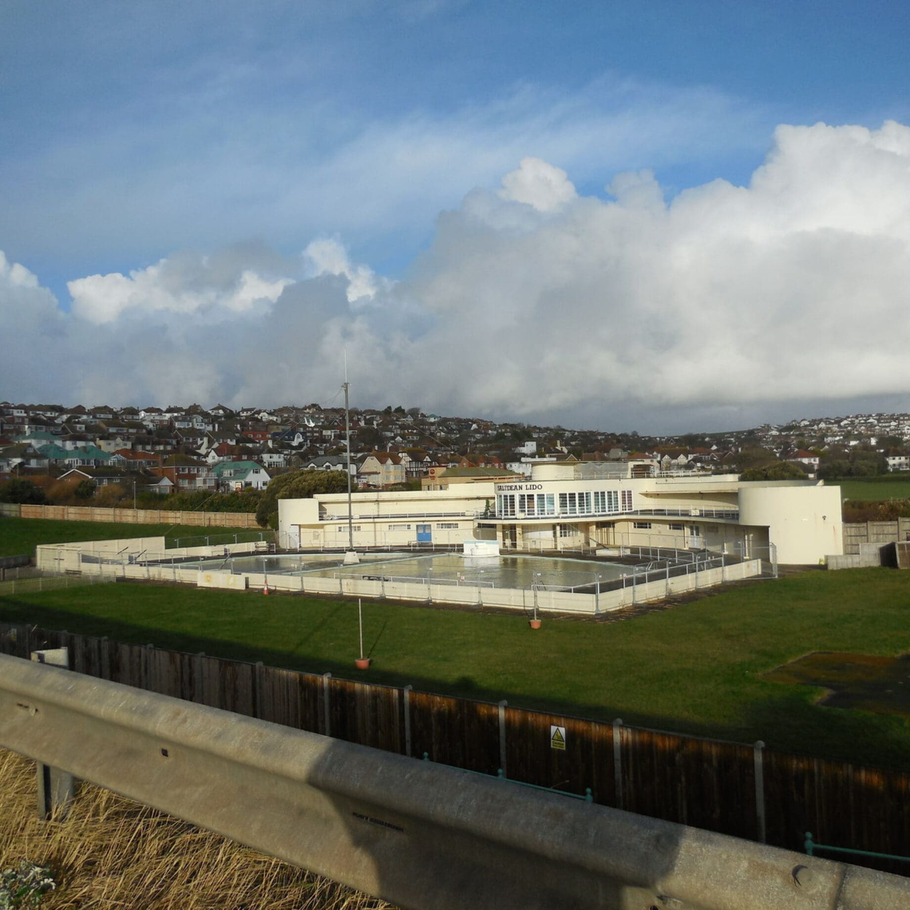 A pale lido building labeled SALTDEAN LIDO sits beside fenced empty outdoor pools on a grassy slope, with terraced houses on the hill and a cloud-filled blue sky.