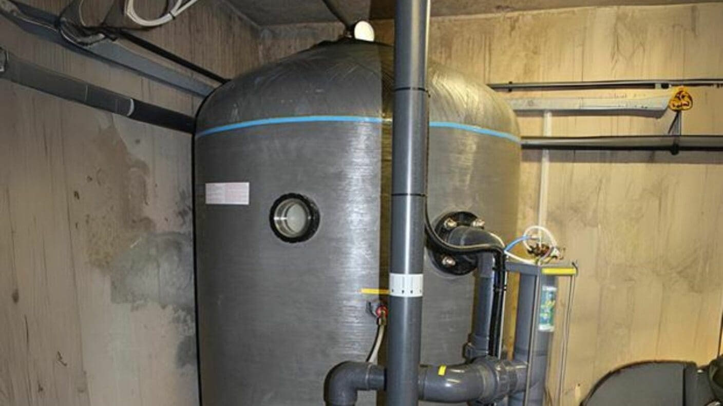 A large vertical gray cylindrical tank with a blue stripe connected to dark PVC pipes and a transparent cartridge filter in a concrete walled utility room.