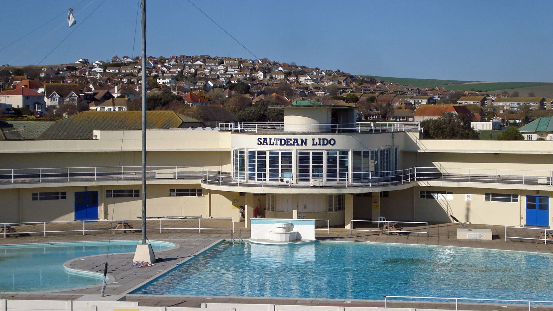A white art-deco building labeled SALTDEAN LIDO fronts a rectangular outdoor swimming pool with clear blue water, a small round pool to the left, and rows of houses on a sunny hillside; visible text: SALTDEAN LIDO, Walls.