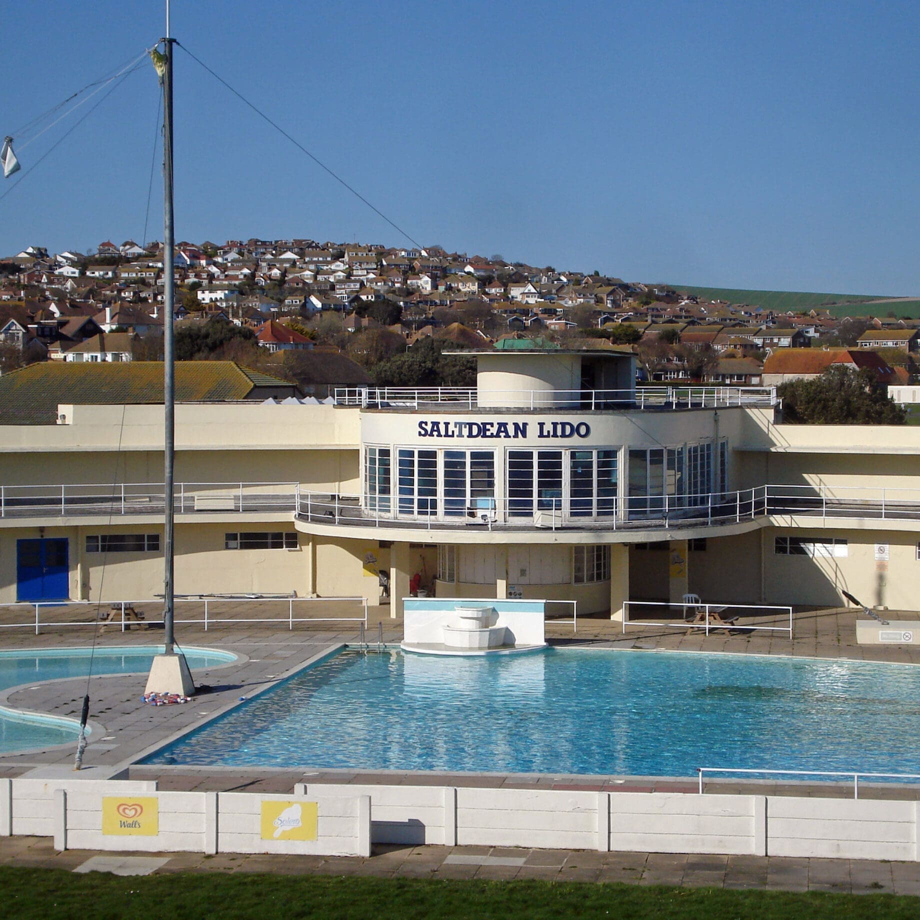 A white art-deco building labeled SALTDEAN LIDO fronts a rectangular outdoor swimming pool with clear blue water, a small round pool to the left, and rows of houses on a sunny hillside; visible text: SALTDEAN LIDO, Walls.