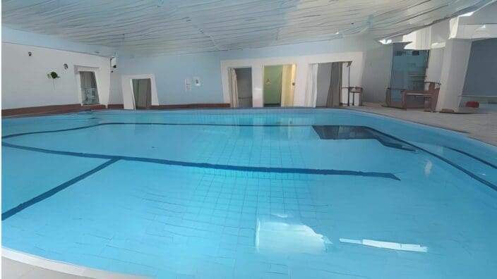 Indoor swimming pool sits calm, water reflecting rippled ceiling; blue-tiled shallow lanes are visible, surrounded by pale walls, doorways, a pool deck with chairs and minimal wall signage.