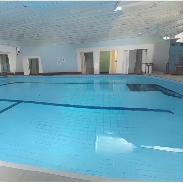 Indoor swimming pool sits calm, water reflecting rippled ceiling; blue-tiled shallow lanes are visible, surrounded by pale walls, doorways, a pool deck with chairs and minimal wall signage.