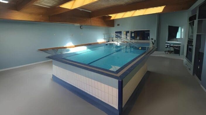 Rectangular indoor pool filled with clear blue water reflecting skylights, with metal handrails and tiled edges, set in a pale-blue room under wooden ceiling beams with an adjacent office window.