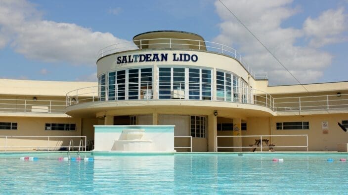 Saltdean Lido building (text: SALTDEAN LIDO) overlooks a calm outdoor pool, showing curved glass windows and white railings under a blue sky on a sunny day.