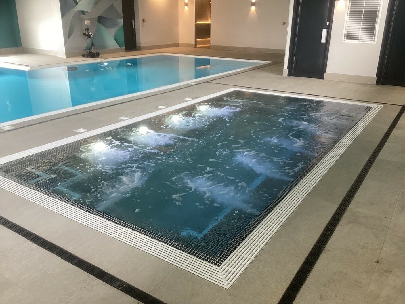 Rectangular indoor whirlpool spa with bubbling, illuminated jets beside a calm lap pool in a tiled pool room with grey floor drains, wall lights, doors and a decorative mural.