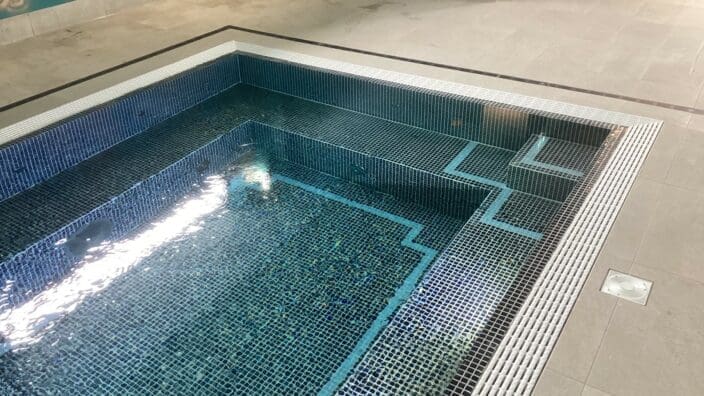Indoor tiled plunge pool shows stepped seating and submerged steps, water reflecting overhead light, surrounded by beige floor tiles and teal-painted walls in a spa-like room