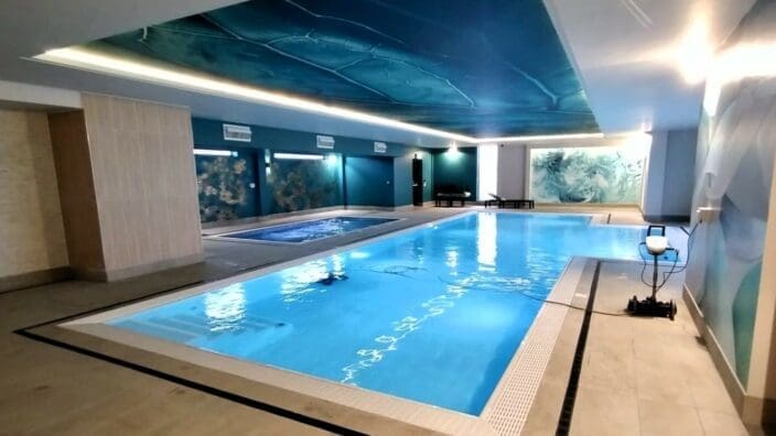 An indoor rectangular pool glows blue, calmly reflecting ceiling lighting while a pool-cleaning robot rests on the deck; the spa room features a muraled teal ceiling, tiled floor and lounge chairs.