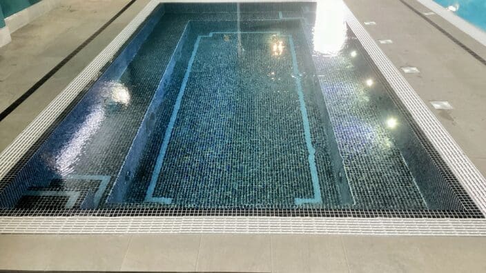 Rectangular tiled spa pool sits recessed in an indoor deck, holding clear water that reflects overhead lights; shallow steps descend into mosaic blue tiles, surrounded by light gray floor tiles.