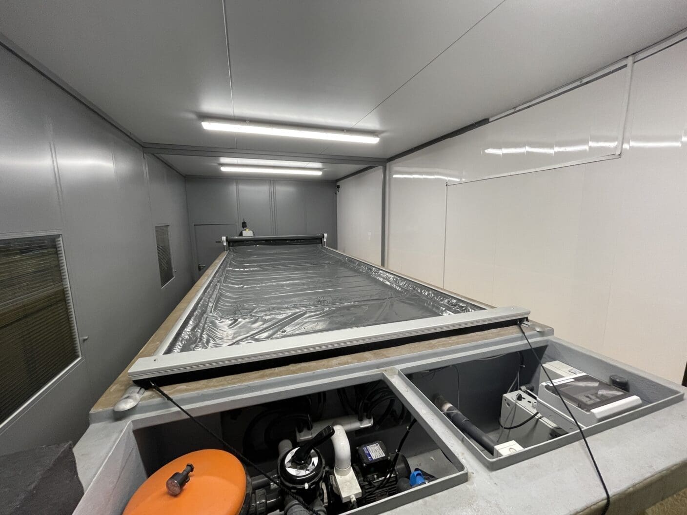 Rectangular industrial basin covered by a grey plastic sheet sits centered, with exposed equipment compartments and piping at the near end, inside a white-gray maintenance room lit by fluorescent lights.