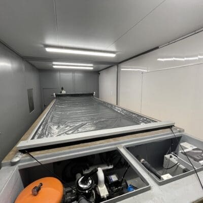 Rectangular industrial basin covered by a grey plastic sheet sits centered, with exposed equipment compartments and piping at the near end, inside a white-gray maintenance room lit by fluorescent lights.