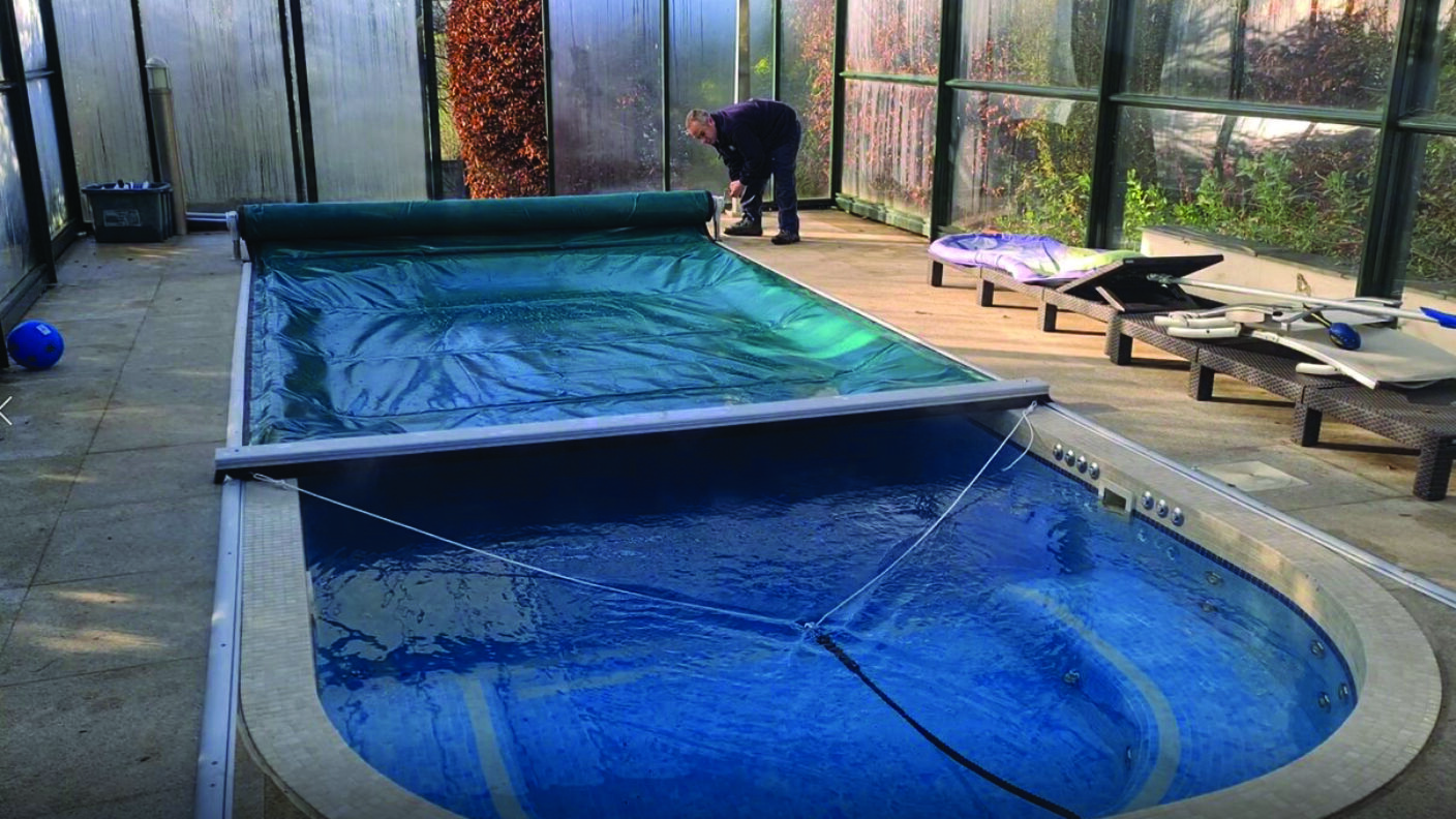 Indoor swimming pool partially covered by a green cover, a person kneeling at the far end adjusting the cover; glass-panel enclosure, deck, lounge chairs and pool equipment along the side.