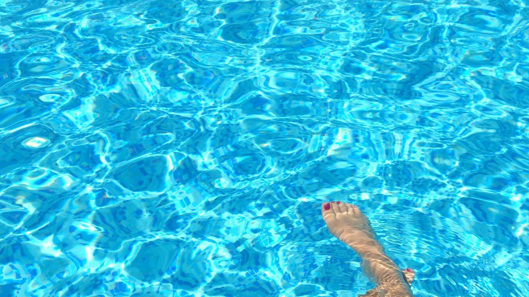 Bare foot with red painted toenails dips into shimmering blue pool water, sending gentle ripples and sunlit reflections across the tiled pool bottom.