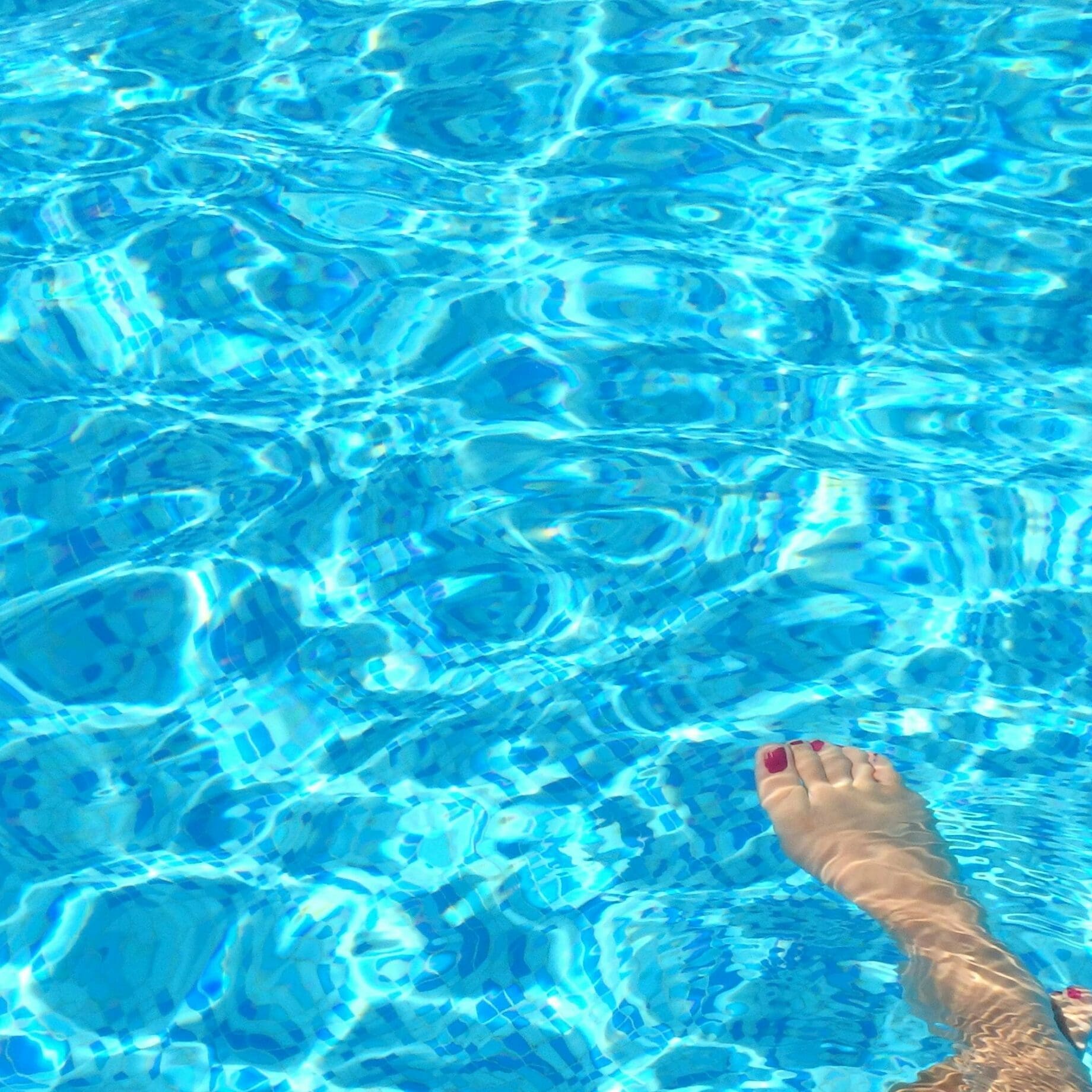 Bare foot with red painted toenails dips into shimmering blue pool water, sending gentle ripples and sunlit reflections across the tiled pool bottom.