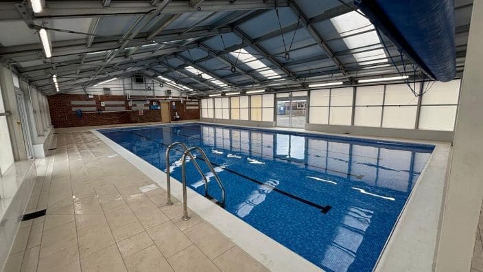 Indoor pool lies calm, ladder at edge reflecting in blue water, in a glass-roofed hall with tiled deck and brick wall; visible text: 1 2 3 4 NO DIVING