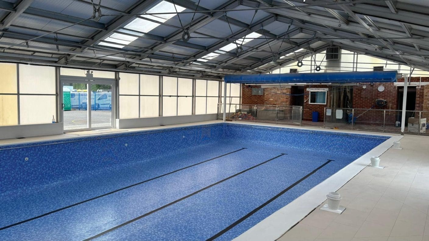 Indoor swimming pool lying empty, lined with blue tiles and black lane markings, inside a glass-and-steel enclosure with equipment and a brick wall; sign reading SHALLOW END 0.9 metres