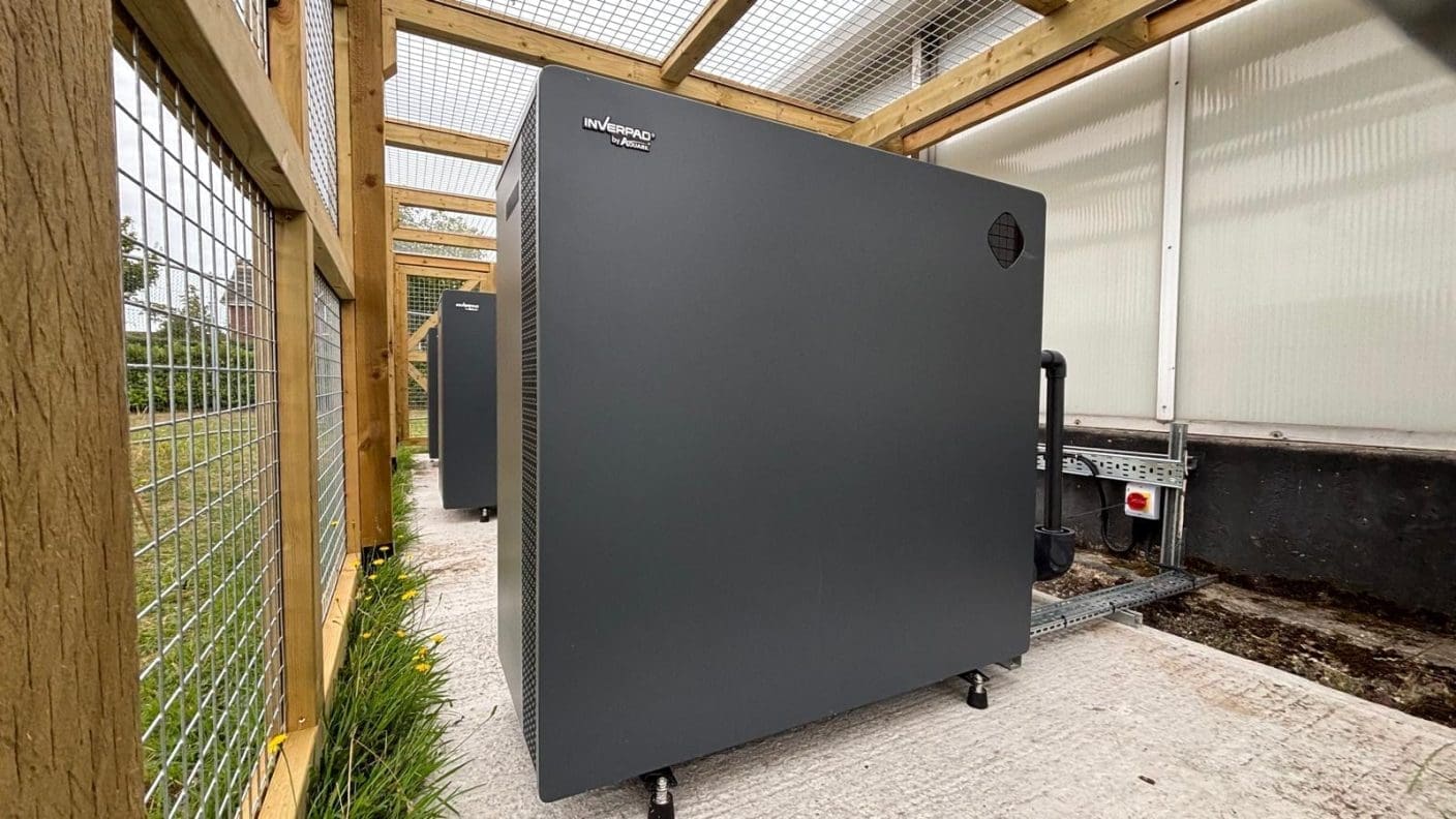 A large dark gray cabinet labeled 'INVERPAD by AUTARK' stands on adjustable feet inside a wooden-framed mesh enclosure, connected to piping and electrical conduit on a concrete pad beside grass and a control switch.