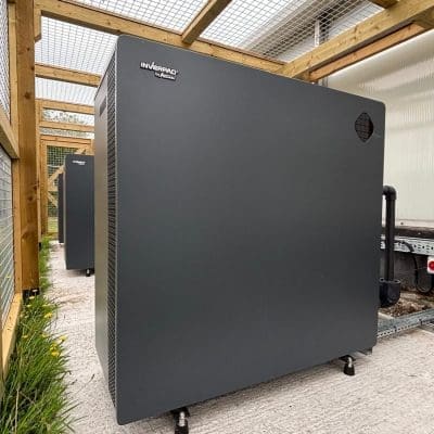 A large dark gray cabinet labeled 'INVERPAD by AUTARK' stands on adjustable feet inside a wooden-framed mesh enclosure, connected to piping and electrical conduit on a concrete pad beside grass and a control switch.