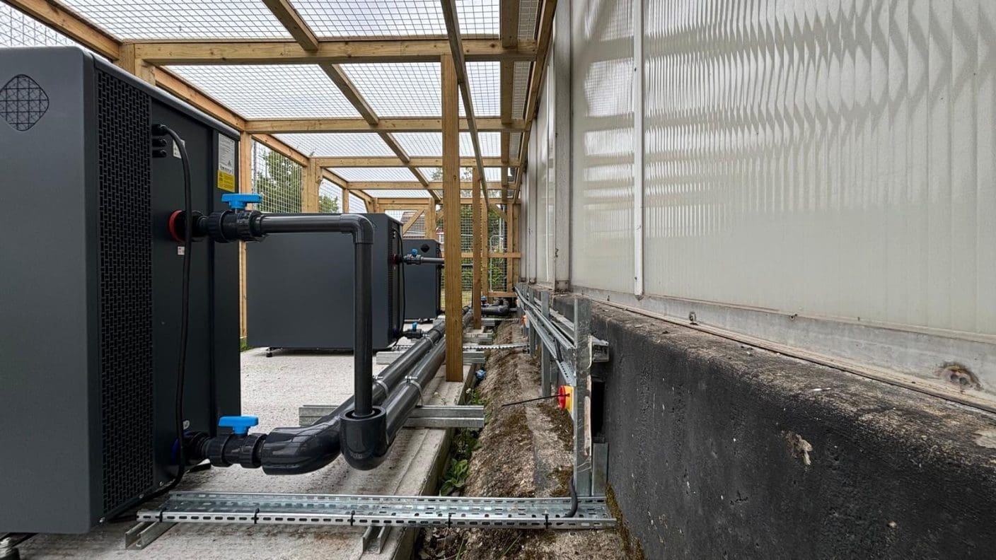 Gray industrial units connected by black PVC pipes and blue-handled valves run along concrete slabs under a wooden lattice canopy beside a translucent greenhouse wall.