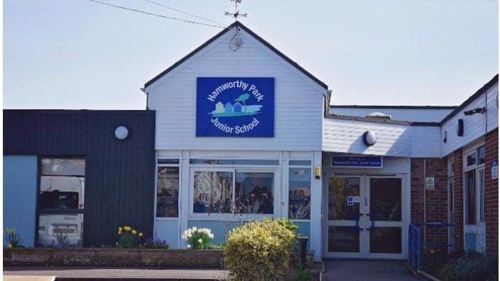 A single-story school building displays a blue sign above the entrance reading "Hamworthy Park Junior School", with glass doors, planters and a paved parking/footpath area under a clear sky.