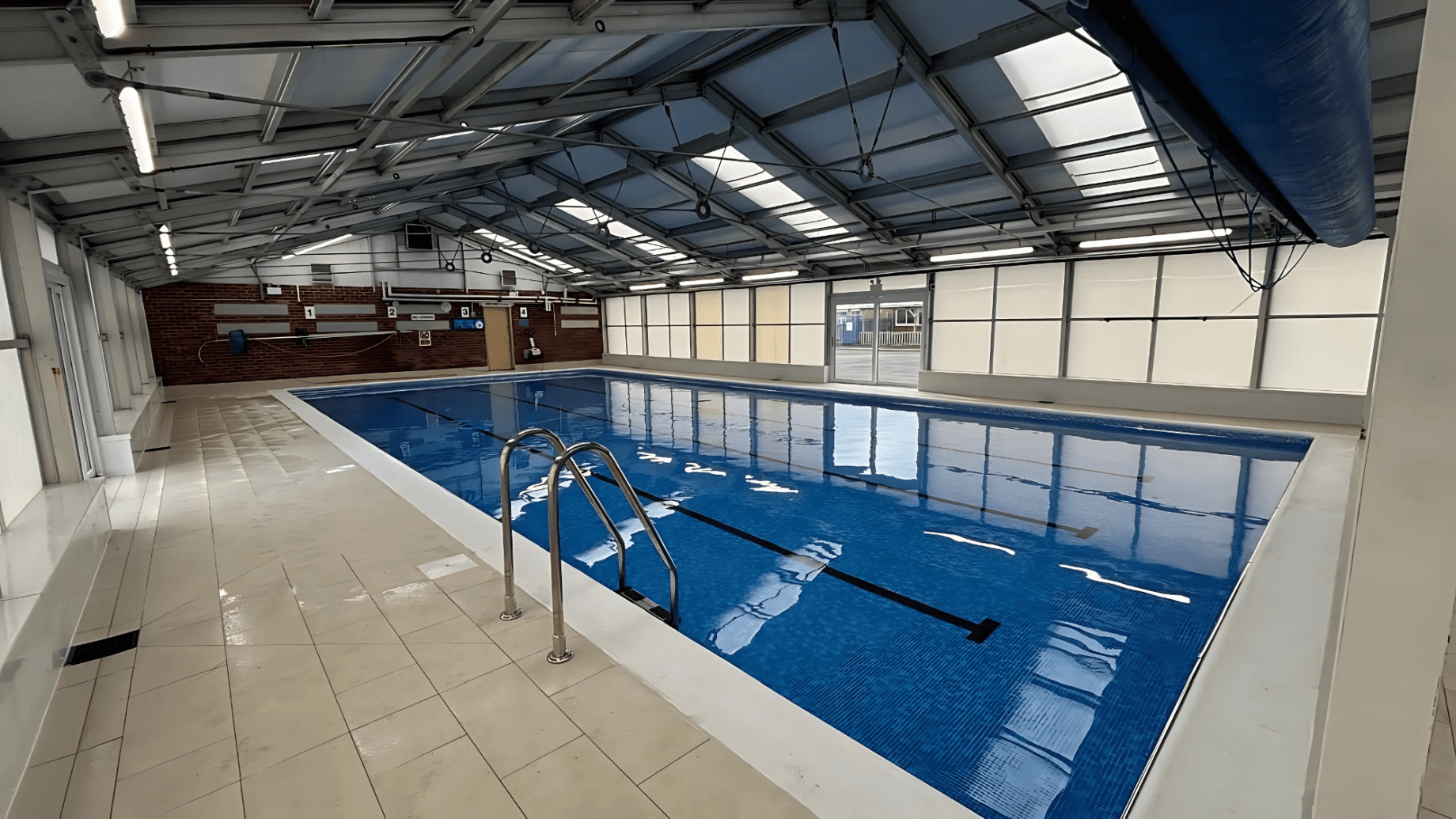 Indoor lap pool sits still, reflecting skylit metal roof; stainless ladder descends into clear blue lanes, tiled deck surrounds, brick far wall displays numbered signs 1 2 3 and pool equipment.