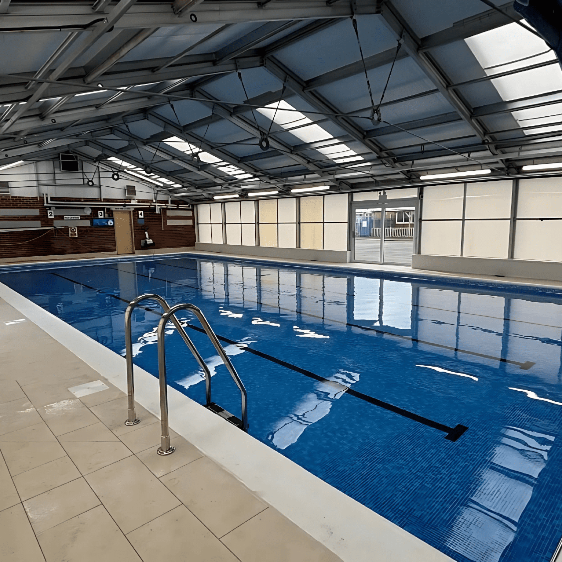 Indoor lap pool sits still, reflecting skylit metal roof; stainless ladder descends into clear blue lanes, tiled deck surrounds, brick far wall displays numbered signs 1 2 3 and pool equipment.