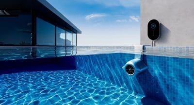 Blue underwater circulation device affixed to tiled pool wall, pumping water and casting rippled light across the pool; a black wall-mounted control unit and modern glass house sit at the rim.