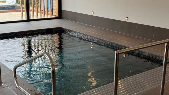 Rectangular indoor pool ripples gently, stainless-steel handrails leading into water, mosaic tile trim and grey tiled deck inside a bright room with recessed wall lights and large glass doors.