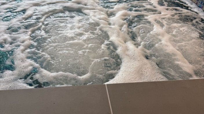 Hot tub basin bubbling vigorously while foam swirls; a stainless square control plate with a blue-centered button rests on the tiled deck beside mosaic pool trim and reflected window light.