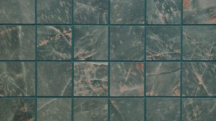 Square green marble tiles form a regular grid, each showing pale pink veining; narrow blue-green grout lines separate them, suggesting a tiled wall or floor surface.
