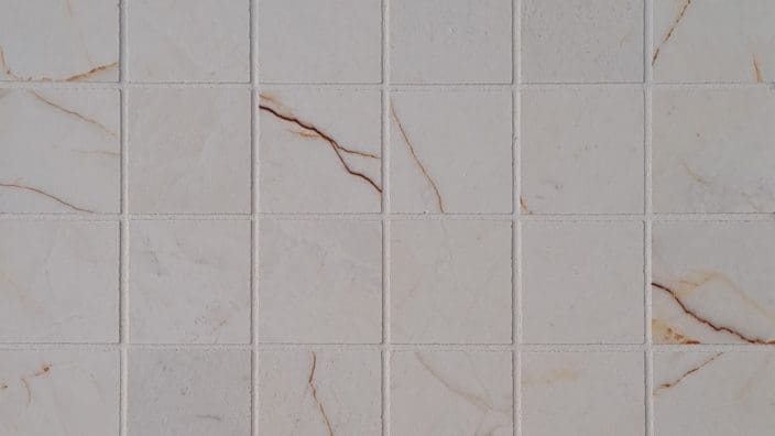 Square white marble tiles arranged in a regular grid, showing thin brown and gold veining across some tiles, separated by narrow grout lines, forming a tiled wall or floor surface.