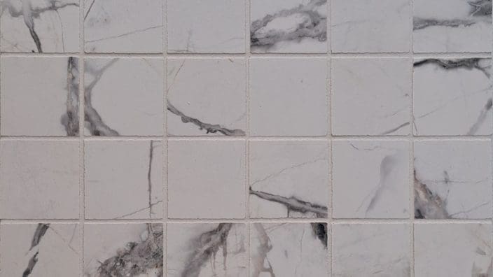 A grid of small square white marble tiles displays gray and black veining, neatly grouted and arranged as a flat wall or floor surface in even rows.