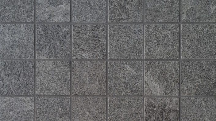 Gray square stone tiles form a regular grid, each rough-textured tile separated by thin grout lines, creating a uniform yet variegated gray surface on a flat floor or wall.